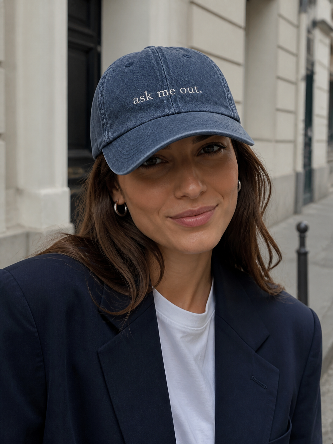 A woman in a charcoal sweatshirt wearing the washed navy 'ask me out.' cap on a city street in front of a stone building.