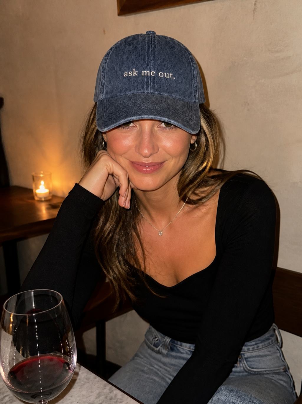 A woman in a white tank top wearing the washed navy 'ask me out.' cap, seated indoors by a window at dusk.