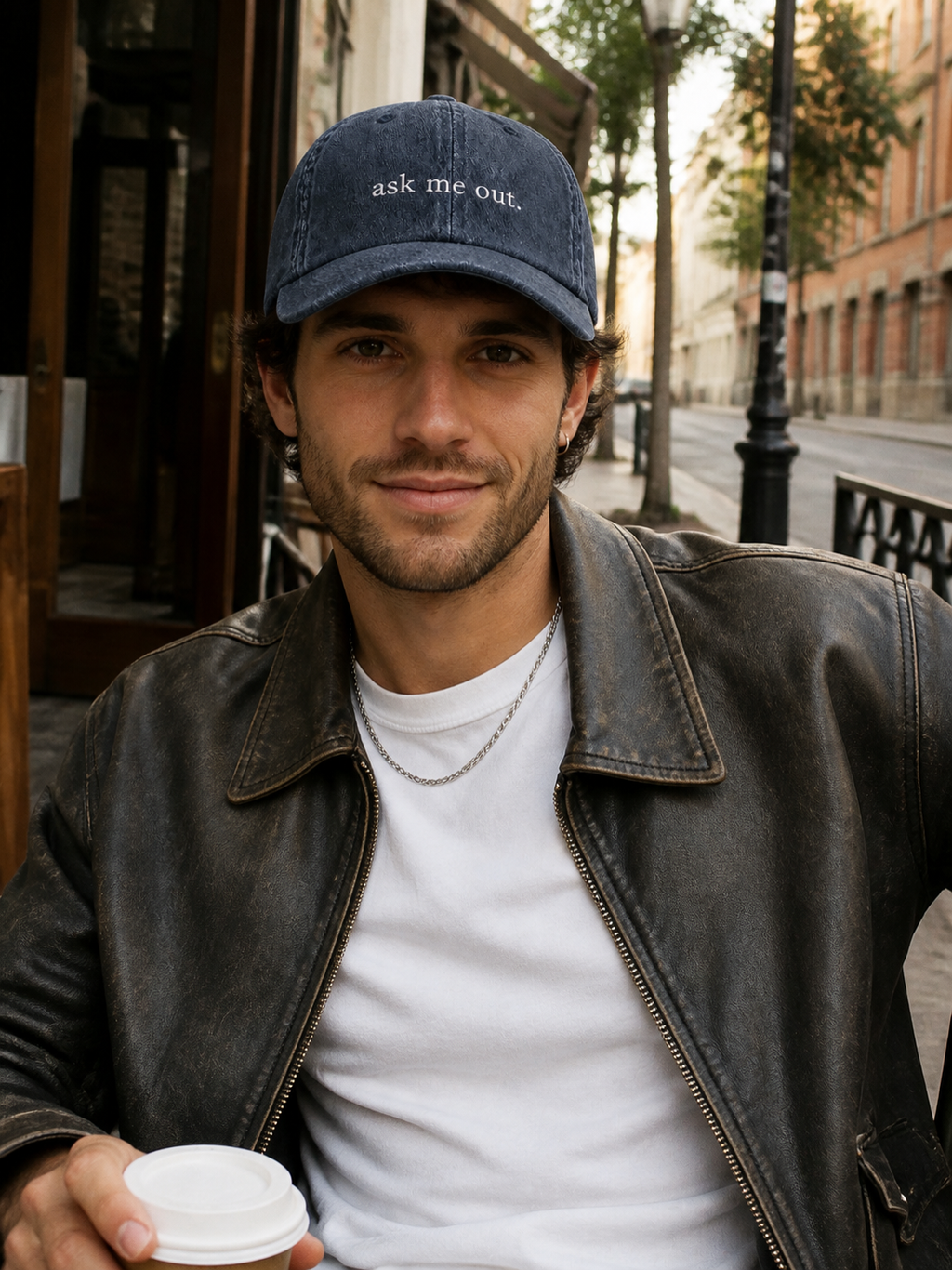 A woman in a black leather jacket holding a coffee cup and wearing the washed navy 'ask me out.' cap at a cafe terrace.