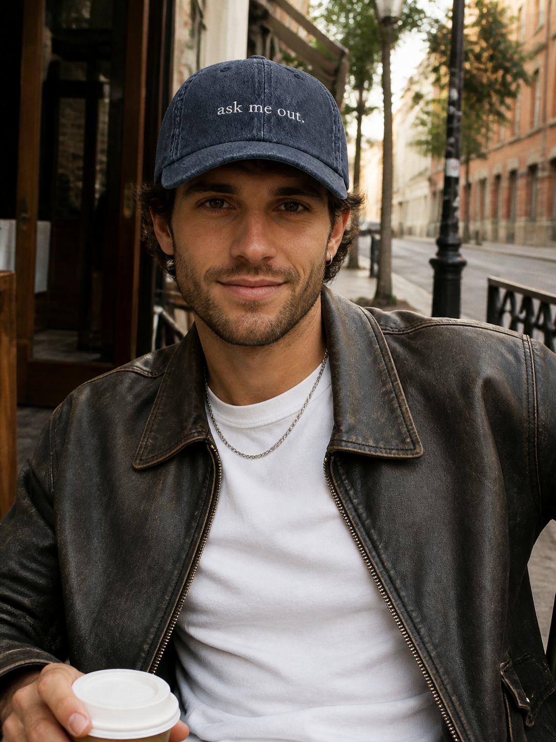 A woman in a black leather jacket holding a coffee cup and wearing the washed navy 'ask me out.' cap at a cafe terrace.