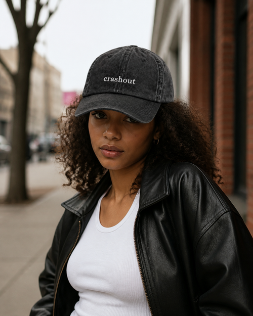 A woman in a black leather jacket and white tank top wearing the washed black 'crashout' cap on a city street.