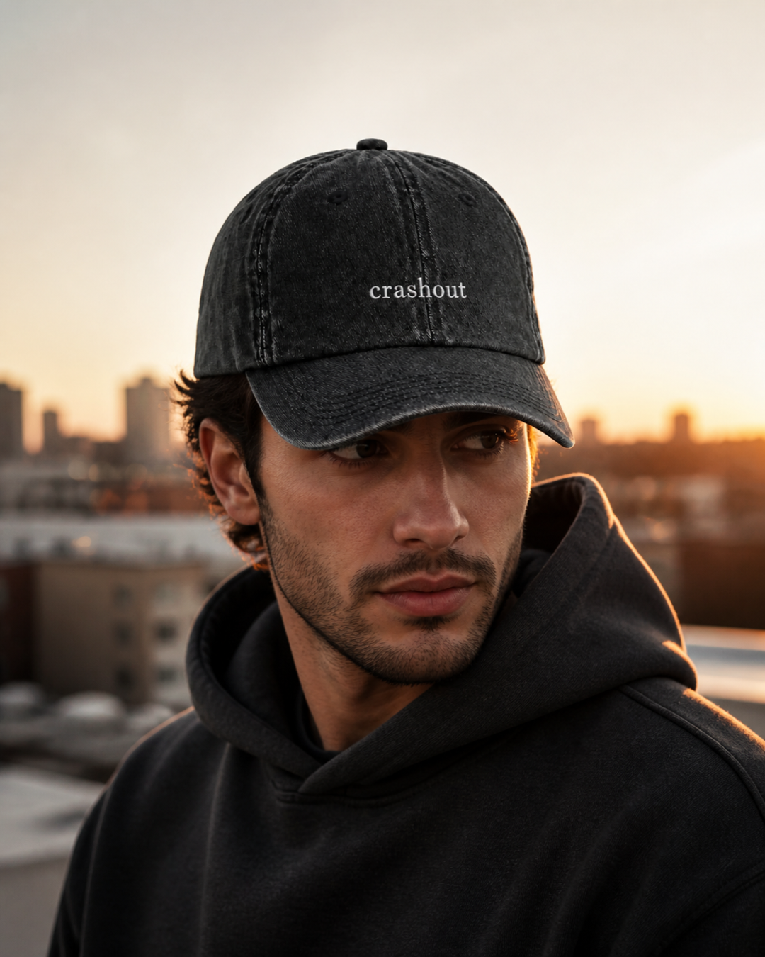 A man in a dark hooded sweatshirt wearing the washed black 'crashout' cap on a rooftop at sunset, city skyline behind him.