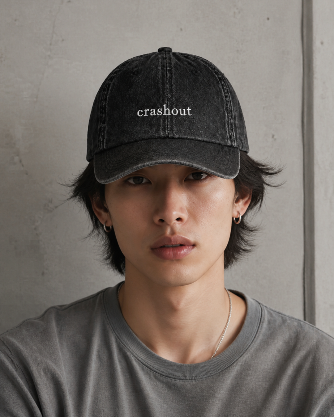 A person in a grey t-shirt wearing the washed black 'crashout' cap against a bare concrete wall.