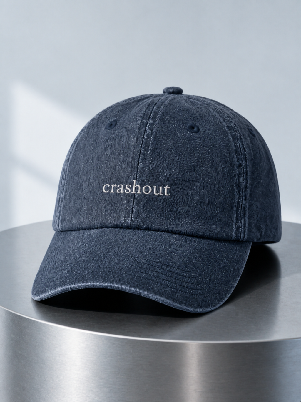 Washed navy 'crashout' cap displayed on a polished steel cylinder plinth in cool daylight.