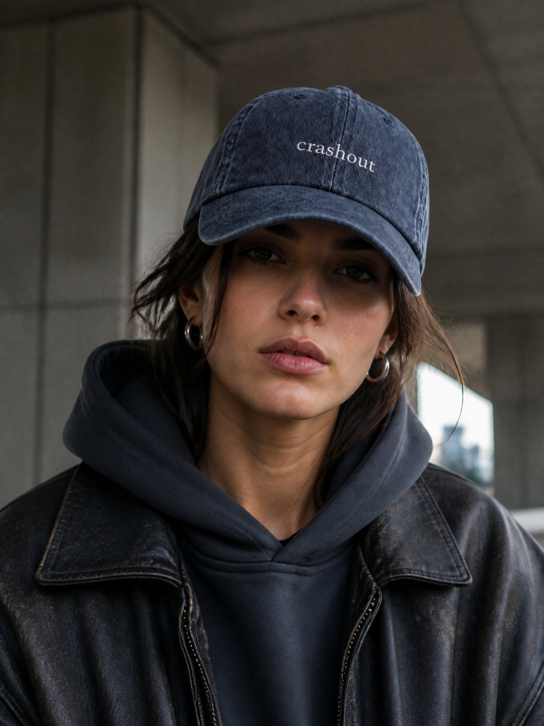 A woman in a charcoal sweatshirt wearing the washed navy 'crashout' cap on a city street in front of a stone building.
