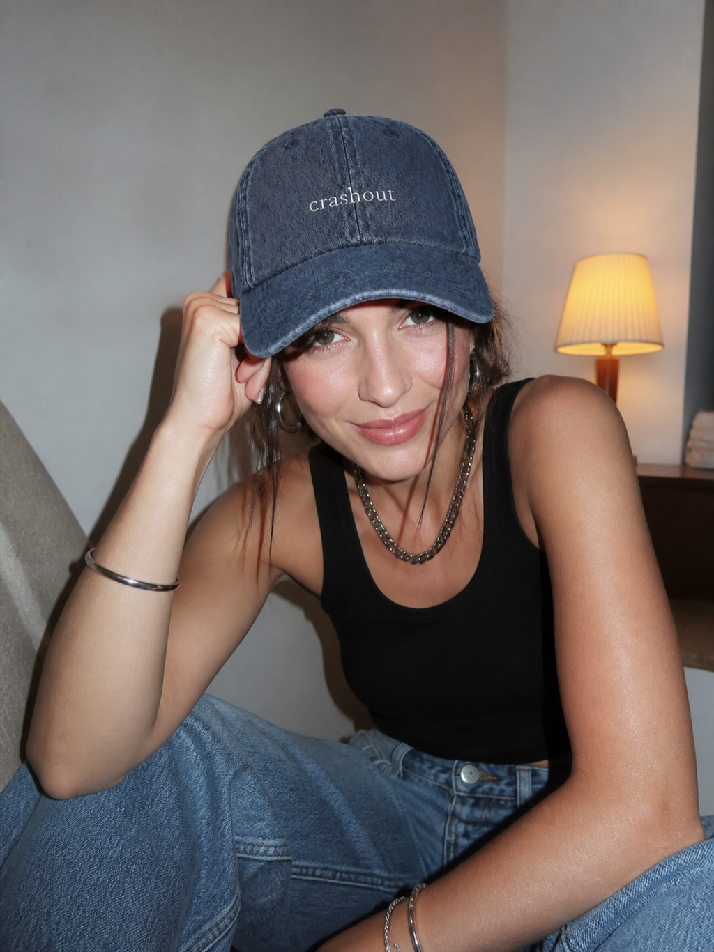 A woman in a white tank top wearing the washed navy 'crashout' cap, seated indoors by a window at dusk.