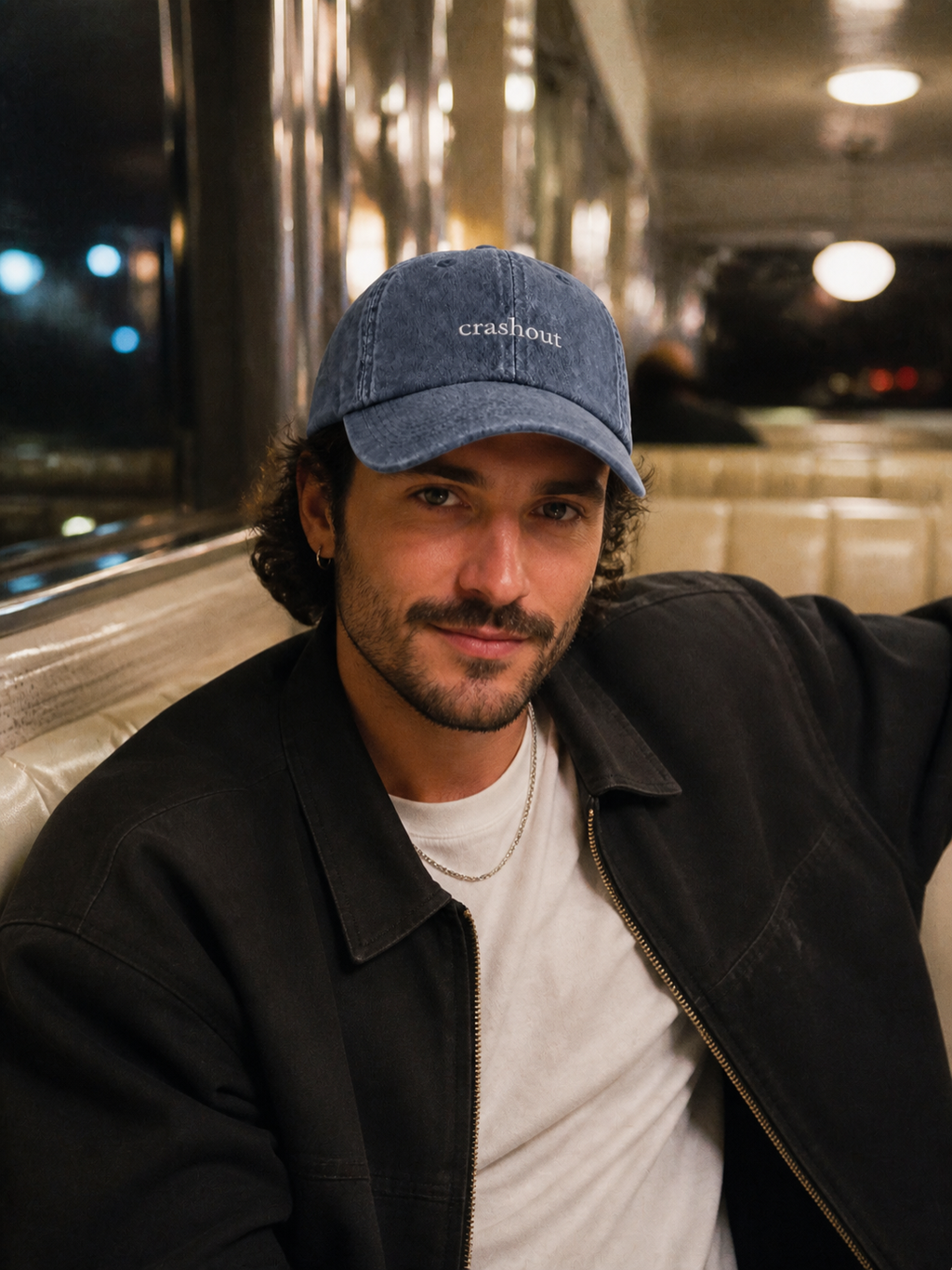 A woman in a black leather jacket holding a coffee cup and wearing the washed navy 'crashout' cap at a cafe terrace.