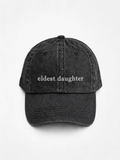 HERO-BLACK Washed black six-panel baseball cap with 'eldest daughter' embroidered in white serif lettering across the front, shown face-on against a plain white background.