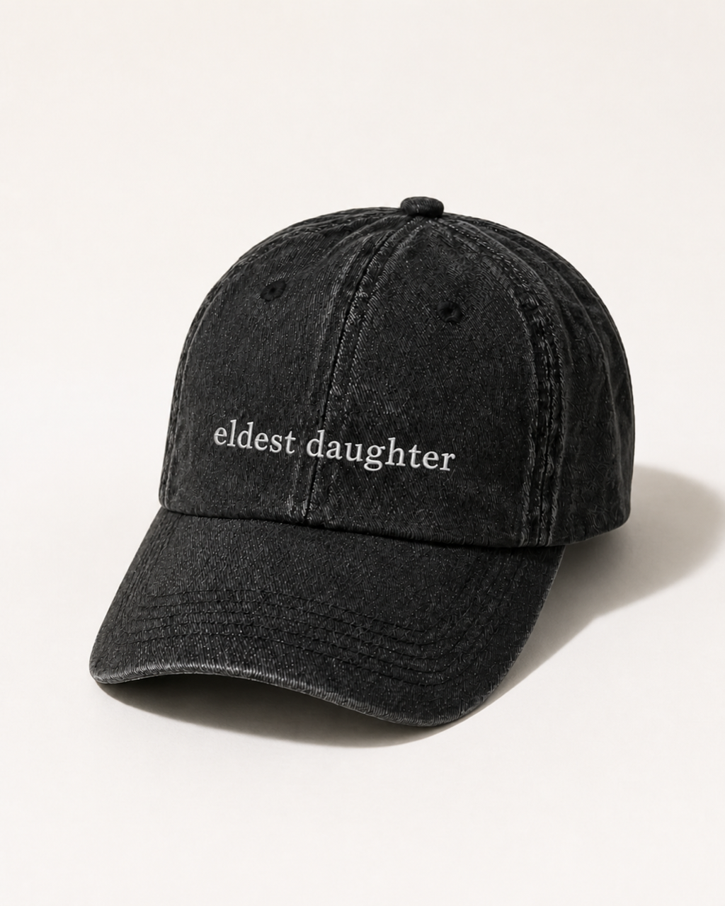 Washed black 'eldest daughter' cap photographed at a three-quarter angle against a warm cream studio backdrop.
