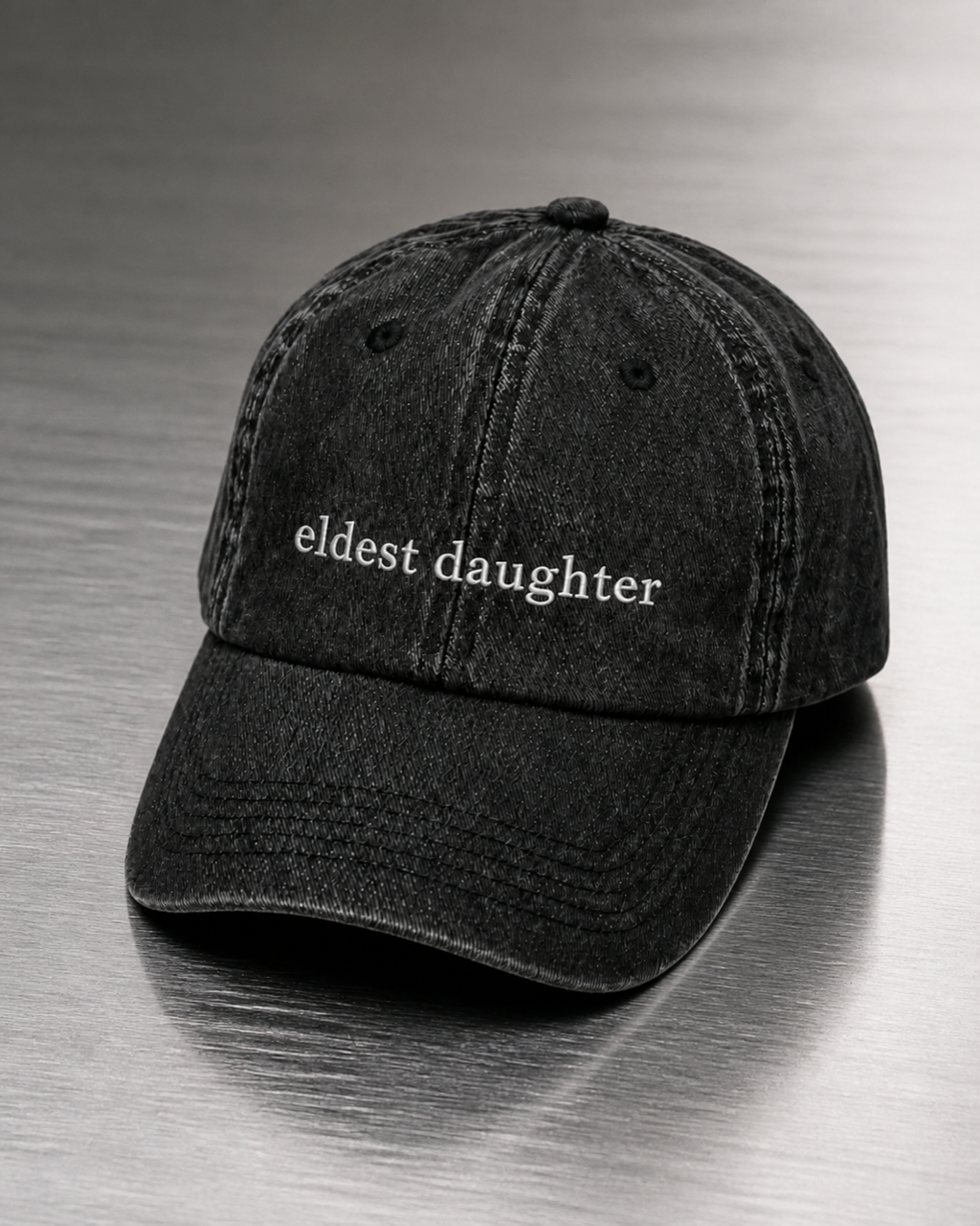 Washed black 'eldest daughter' cap resting on a brushed steel surface under cool directional light.