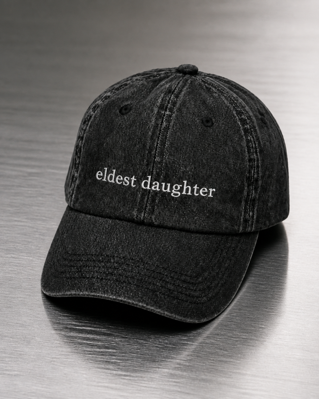 Washed black 'eldest daughter' cap resting on a brushed steel surface under cool directional light.