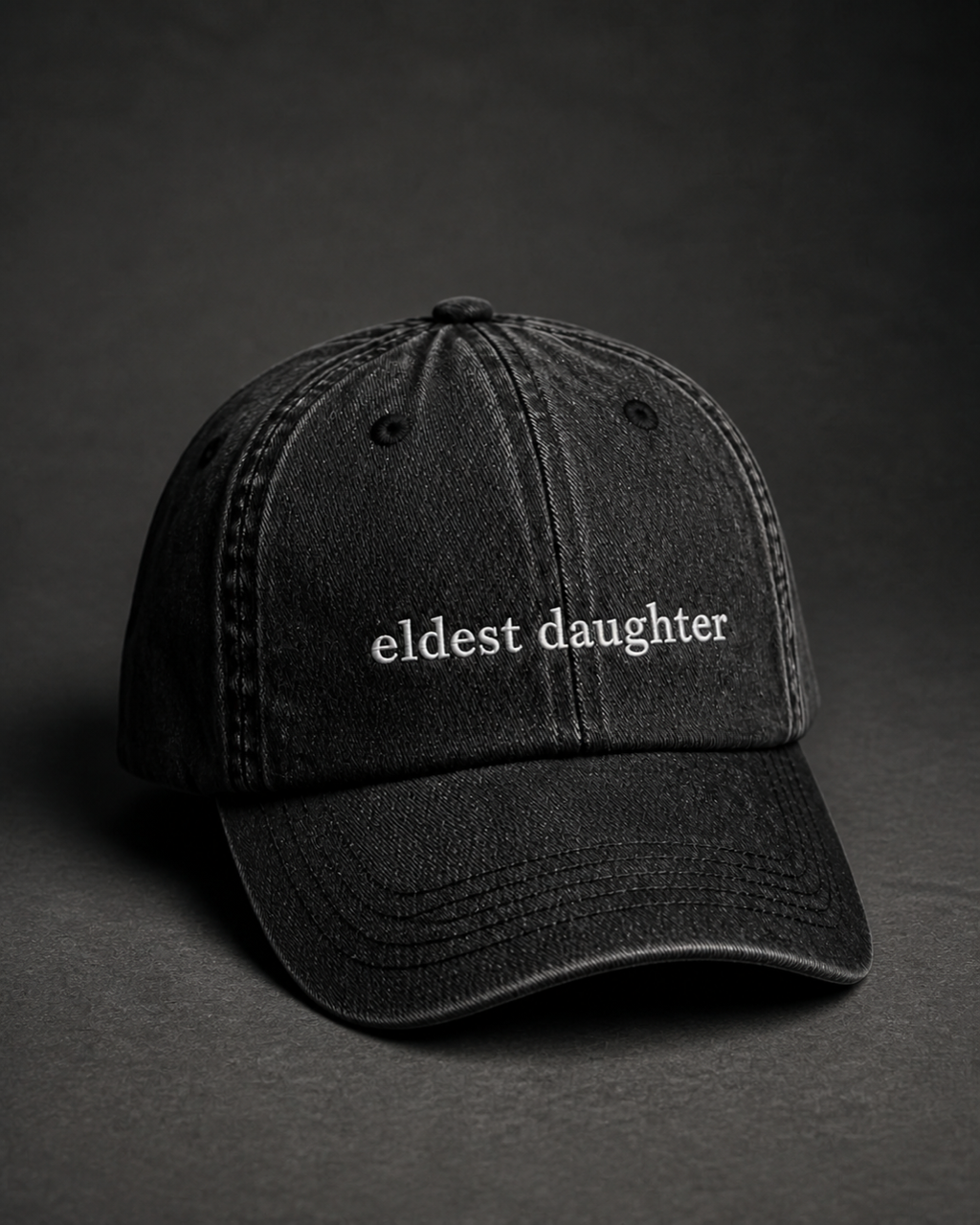 Washed black 'eldest daughter' cap on a dark charcoal backdrop, lit from the side to pick out the embroidery.