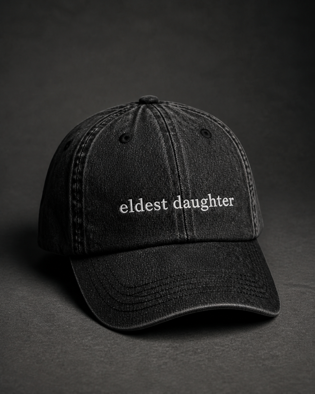 Washed black 'eldest daughter' cap on a dark charcoal backdrop, lit from the side to pick out the embroidery.