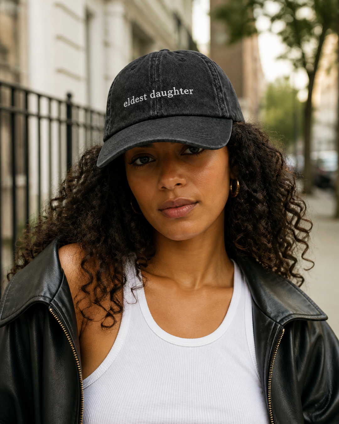 A woman in a black leather jacket and white tank top wearing the washed black 'eldest daughter' cap on a city street.