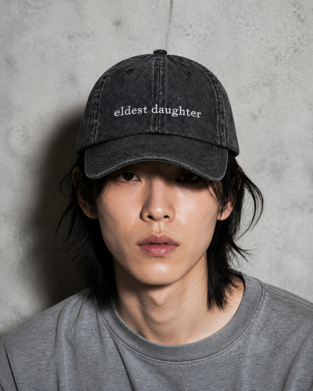 A person in a grey t-shirt wearing the washed black 'eldest daughter' cap against a bare concrete wall.