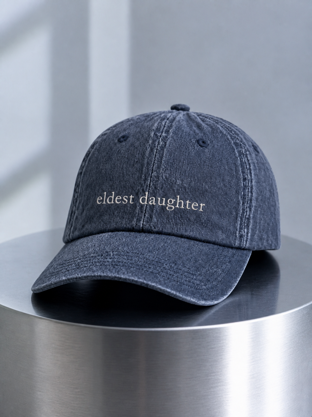 Washed navy 'eldest daughter' cap displayed on a polished steel cylinder plinth in cool daylight.