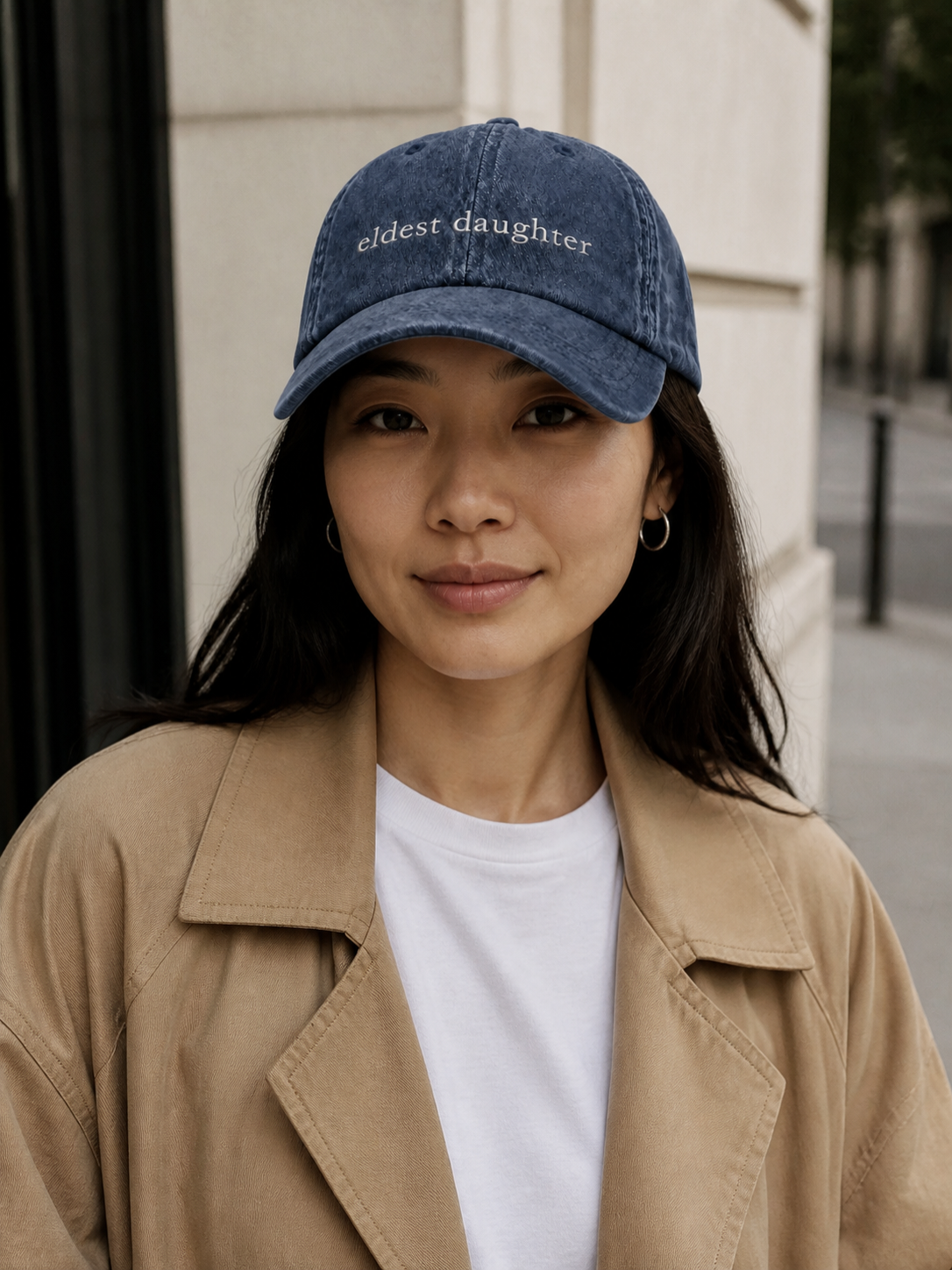 A woman in a charcoal sweatshirt wearing the washed navy 'eldest daughter' cap on a city street in front of a stone building.
