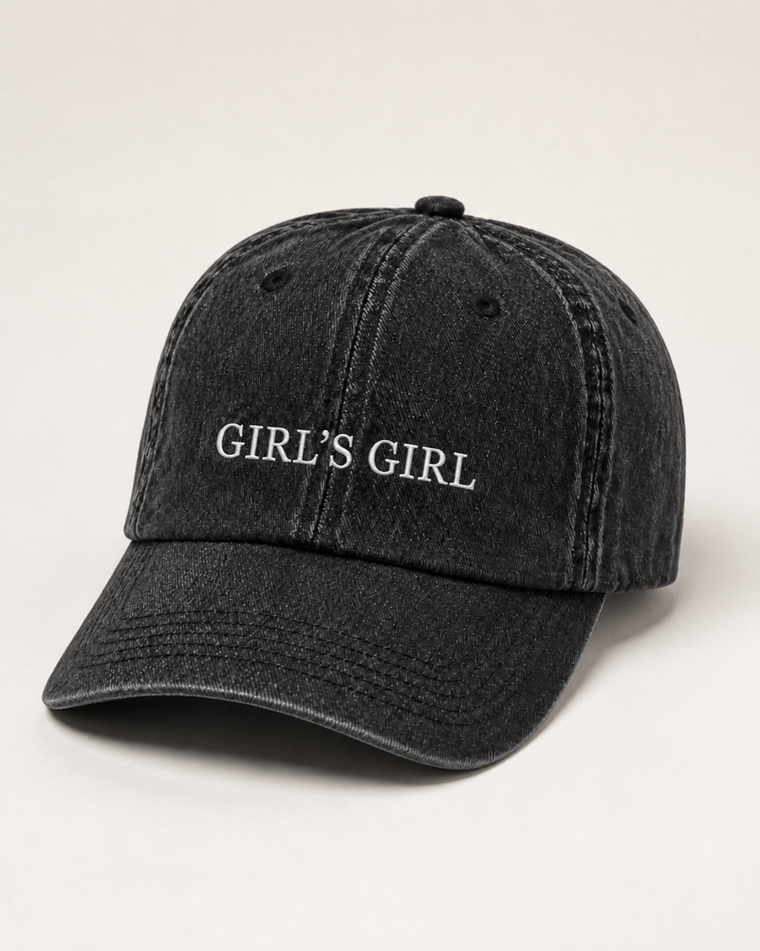 Washed black 'GIRL'S GIRL' cap photographed at a three-quarter angle against a warm cream studio backdrop.