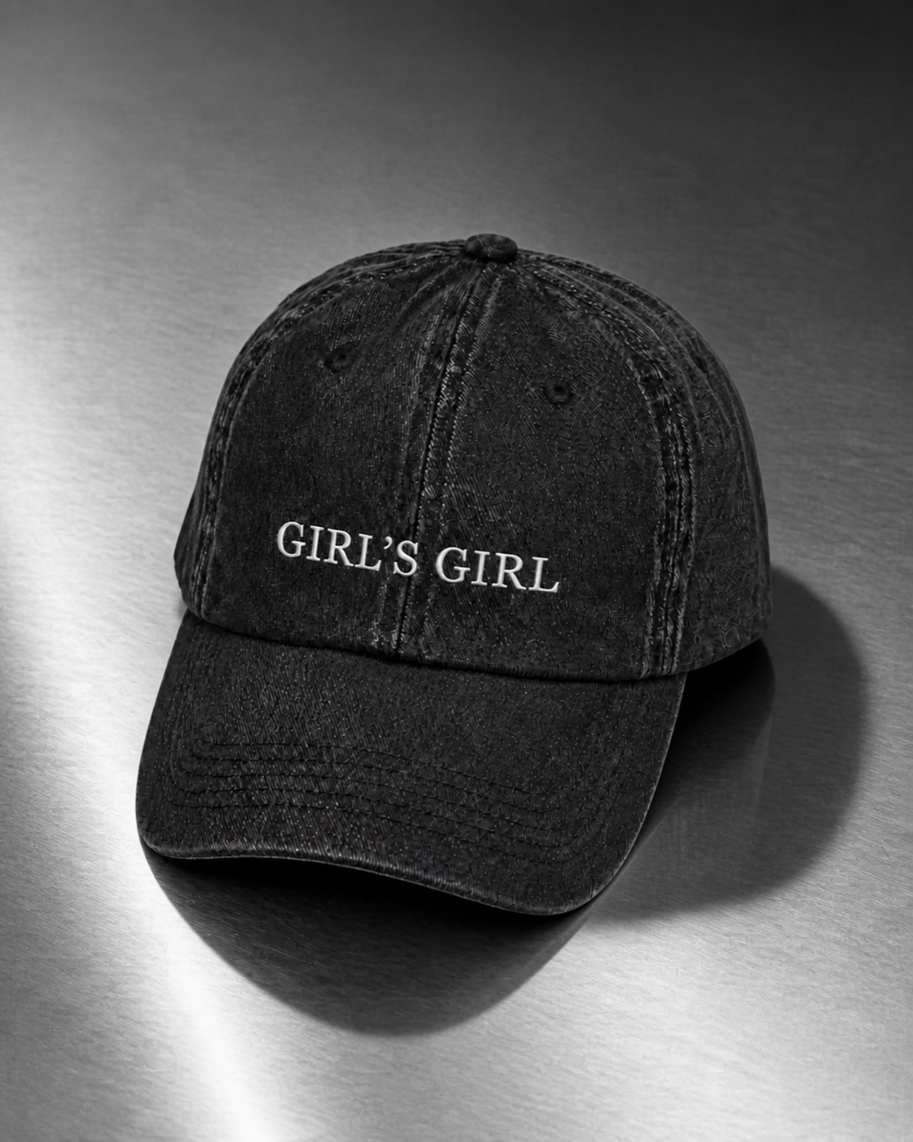 Washed black 'GIRL'S GIRL' cap resting on a brushed steel surface under cool directional light.