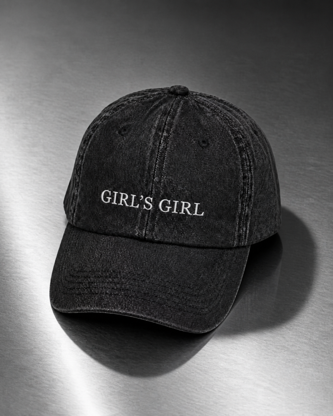 Washed black 'GIRL'S GIRL' cap resting on a brushed steel surface under cool directional light.