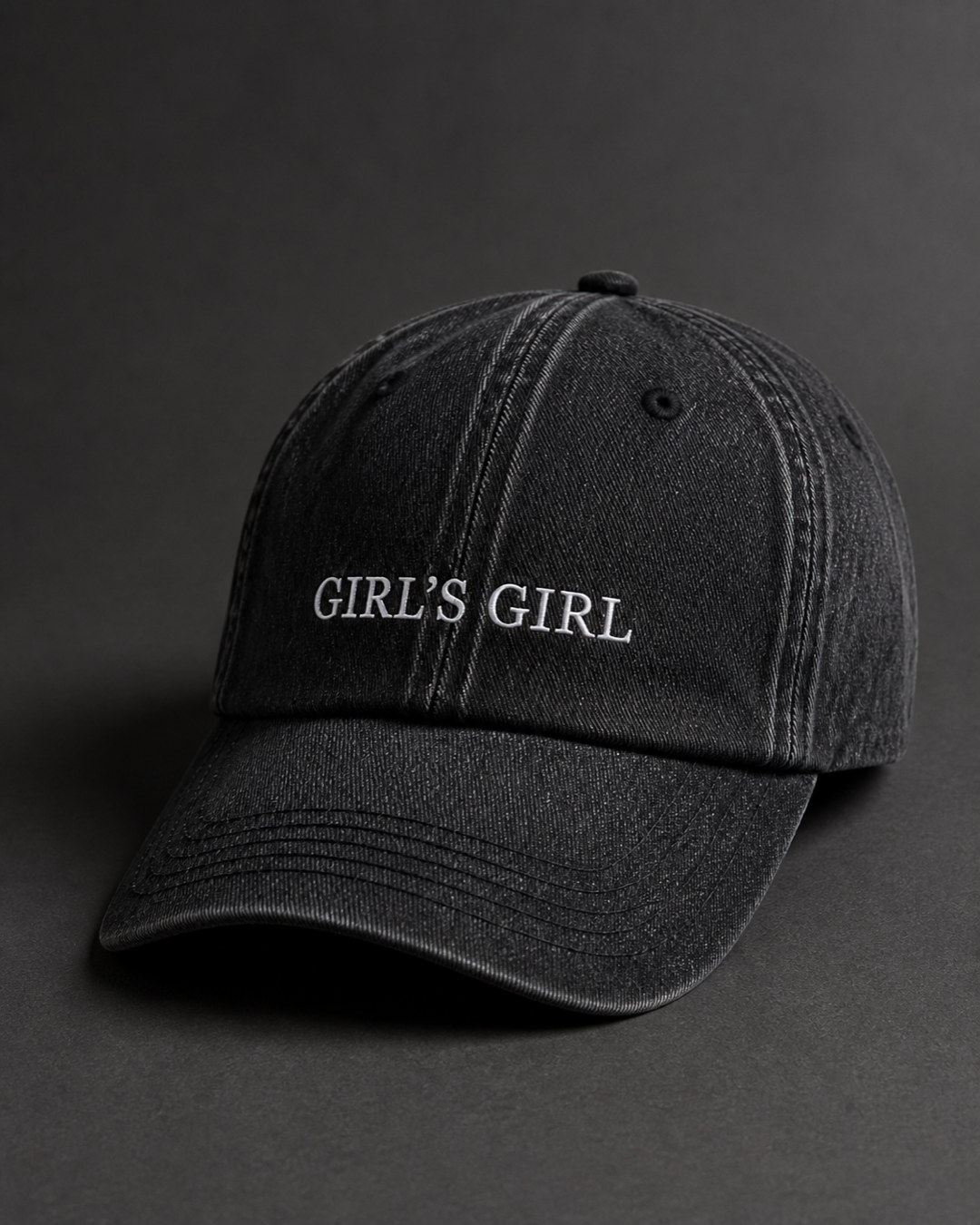 Washed black 'GIRL'S GIRL' cap on a dark charcoal backdrop, lit from the side to pick out the embroidery.