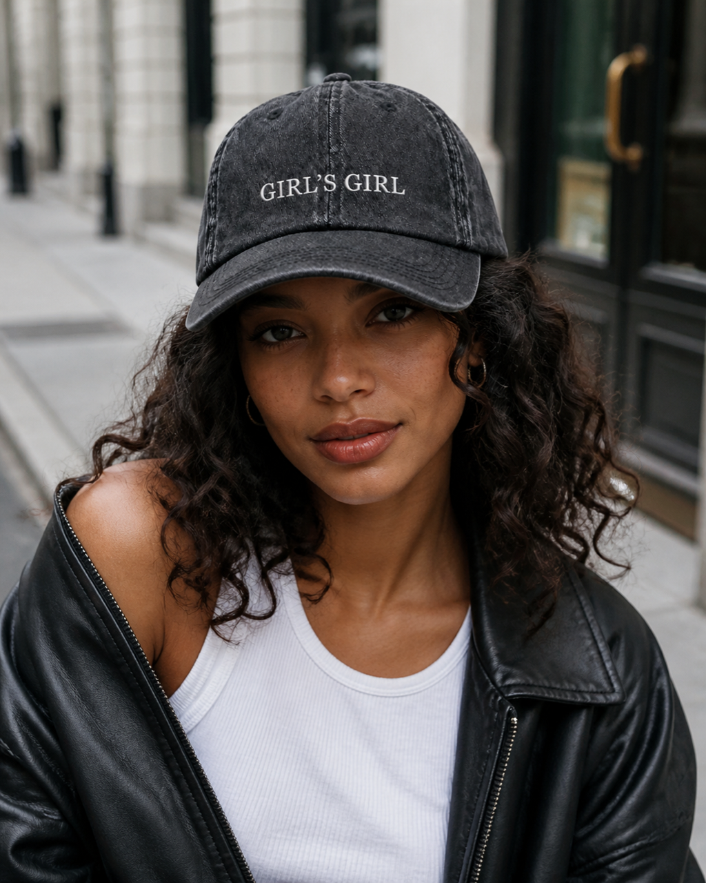 A woman in a black leather jacket and white tank top wearing the washed black 'GIRL'S GIRL' cap on a city street.