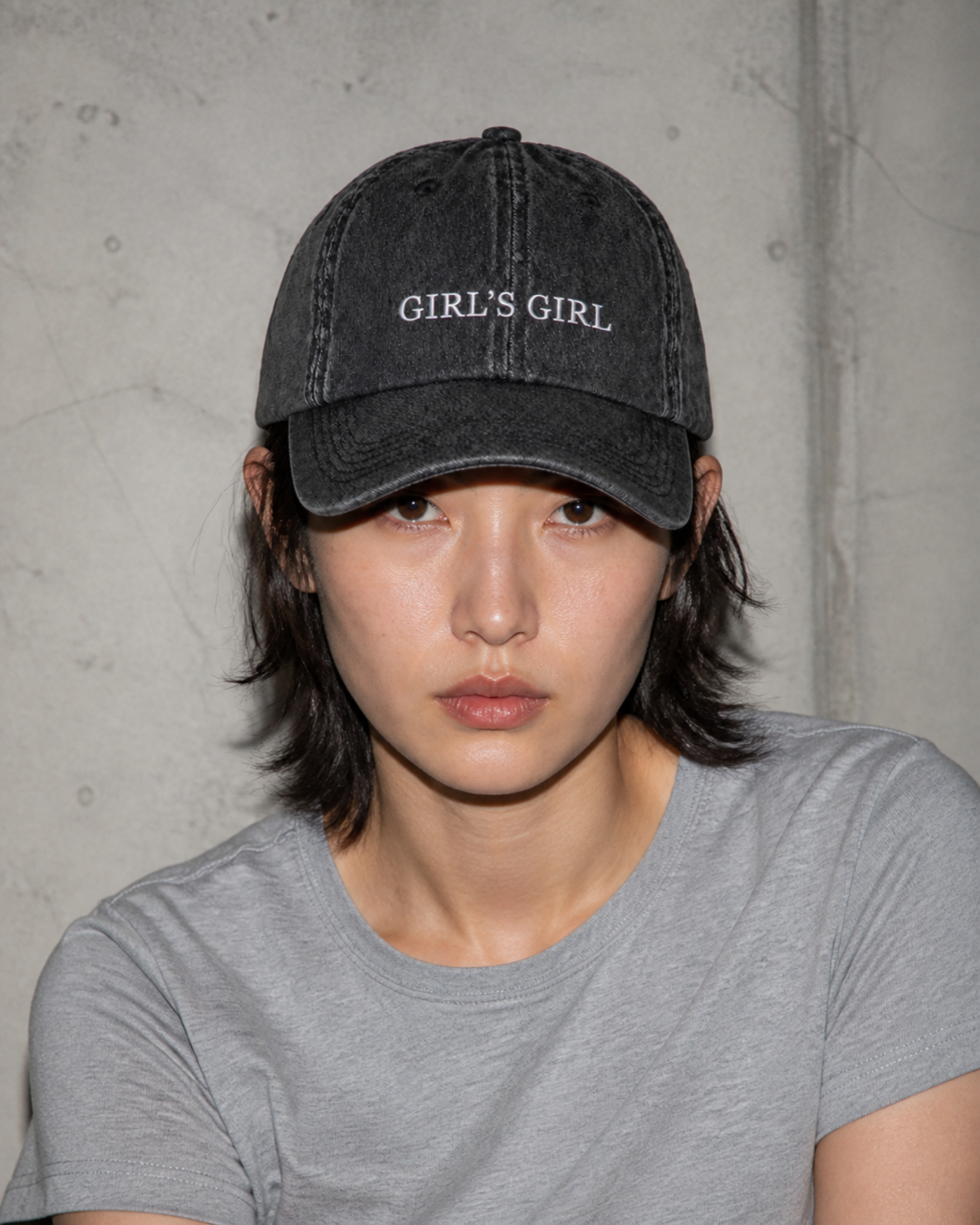 A person in a grey t-shirt wearing the washed black 'GIRL'S GIRL' cap against a bare concrete wall.