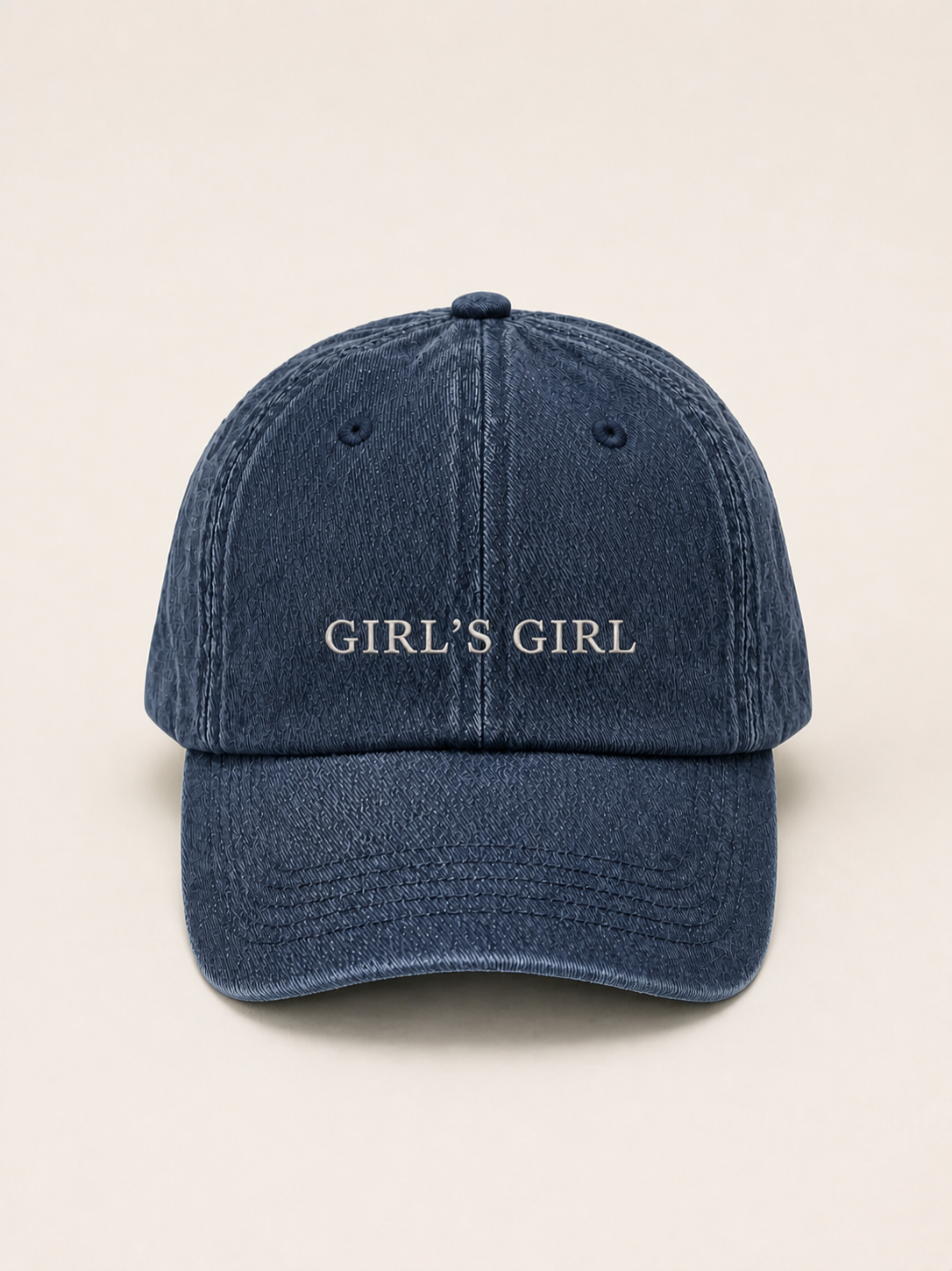 Washed navy 'GIRL'S GIRL' cap photographed face-on against a warm cream studio backdrop.