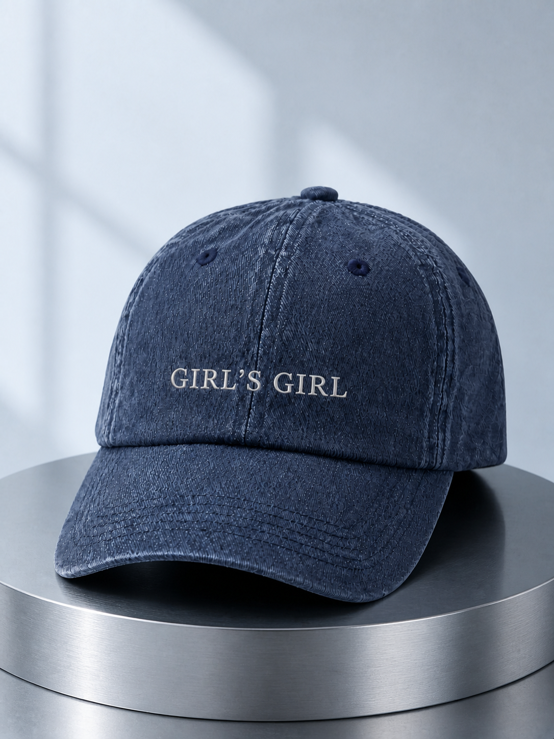 Washed navy 'GIRL'S GIRL' cap displayed on a polished steel cylinder plinth in cool daylight.