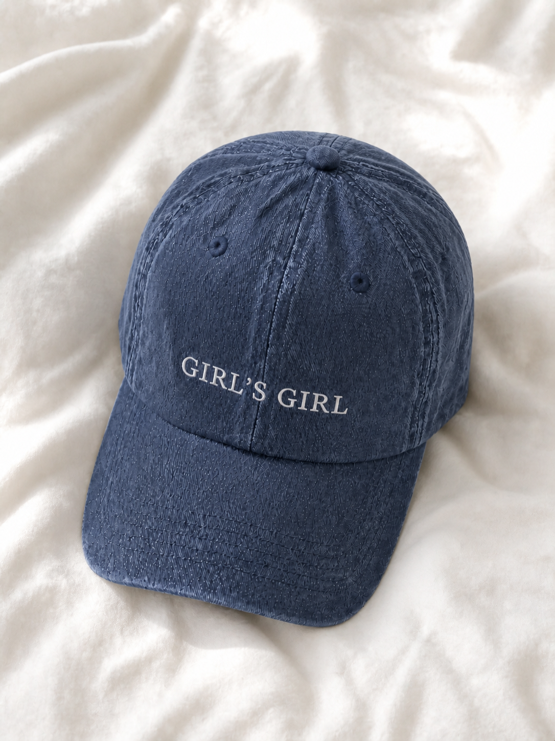 Washed navy 'GIRL'S GIRL' cap resting on rumpled cream linen.