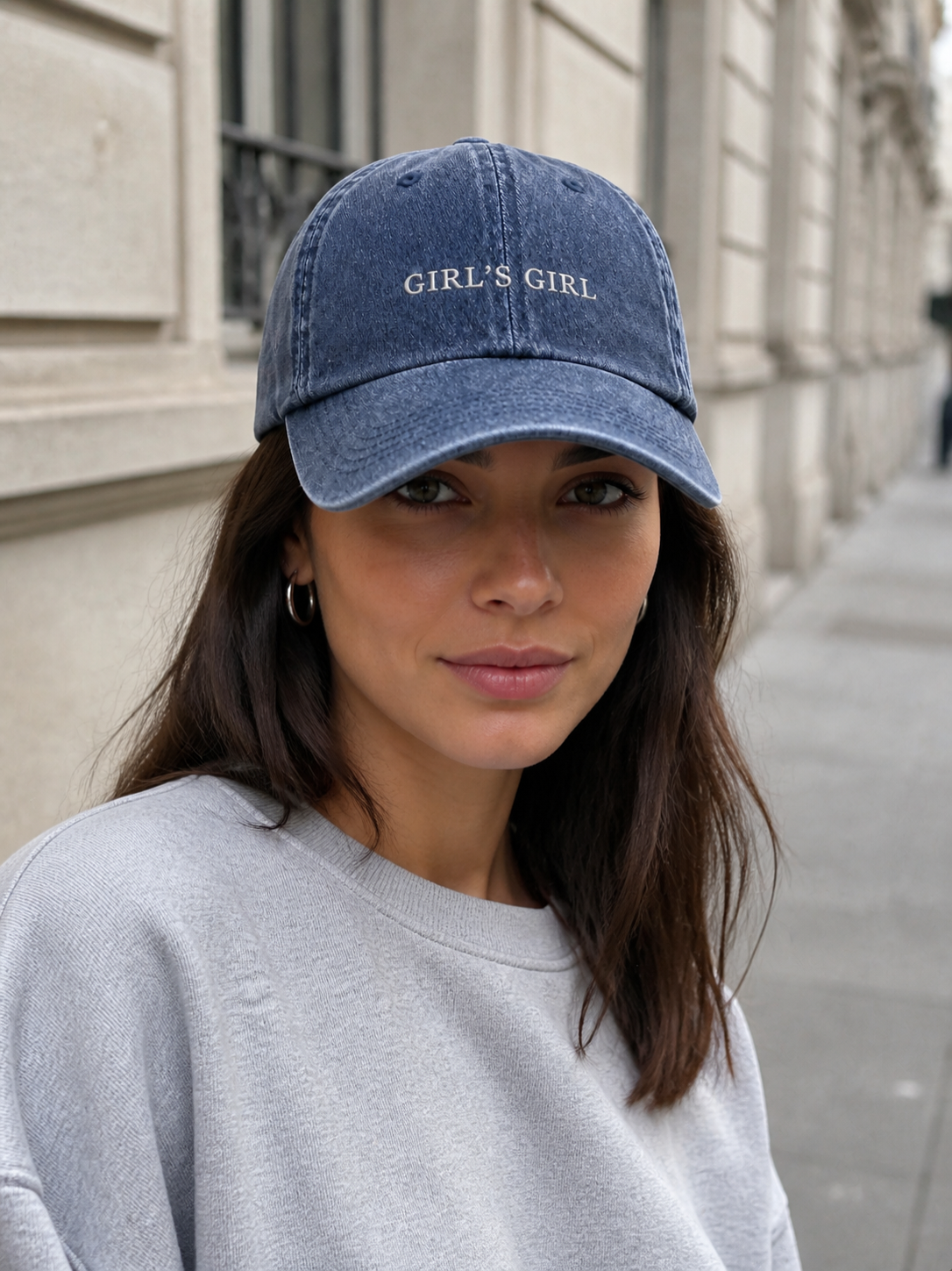 A woman in a charcoal sweatshirt wearing the washed navy 'GIRL'S GIRL' cap on a city street in front of a stone building.