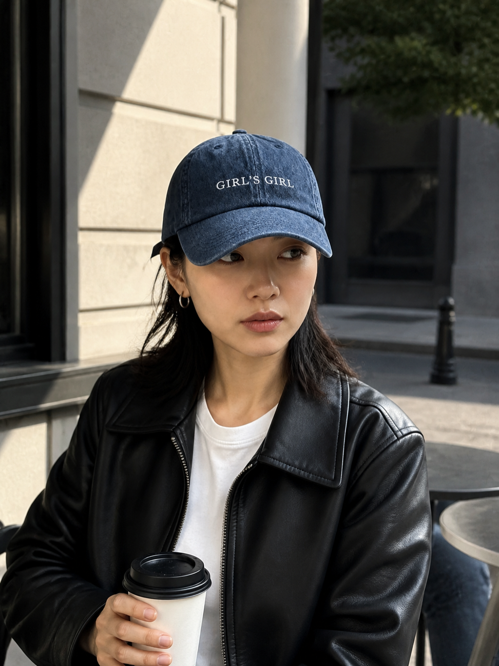 A woman in a black leather jacket holding a coffee cup and wearing the washed navy 'GIRL'S GIRL' cap at a cafe terrace.