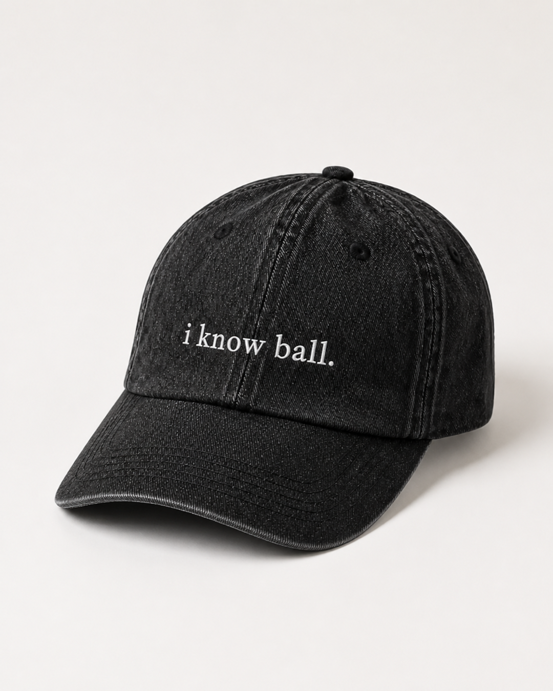 Washed black 'i know ball.' cap photographed at a three-quarter angle against a warm cream studio backdrop.