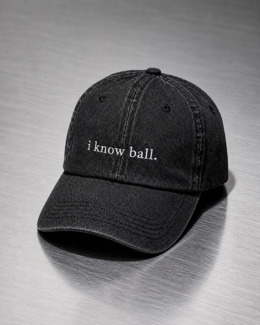 Washed black 'i know ball.' cap resting on a brushed steel surface under cool directional light.