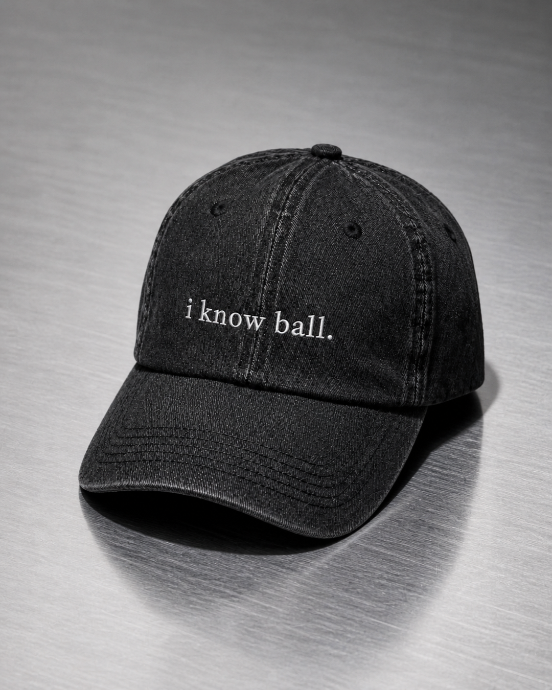 Washed black 'i know ball.' cap resting on a brushed steel surface under cool directional light.