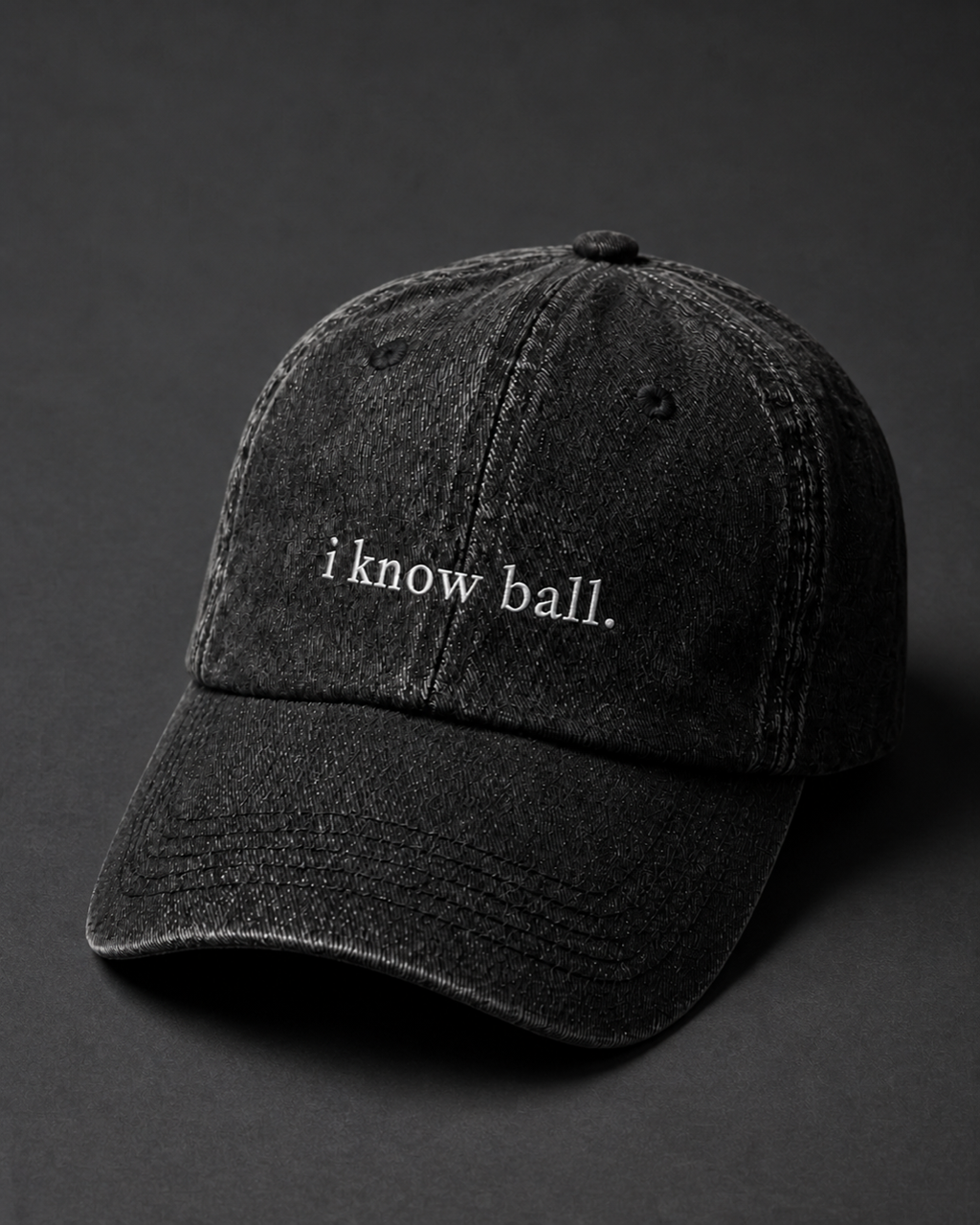 Washed black 'i know ball.' cap on a dark charcoal backdrop, lit from the side to pick out the embroidery.