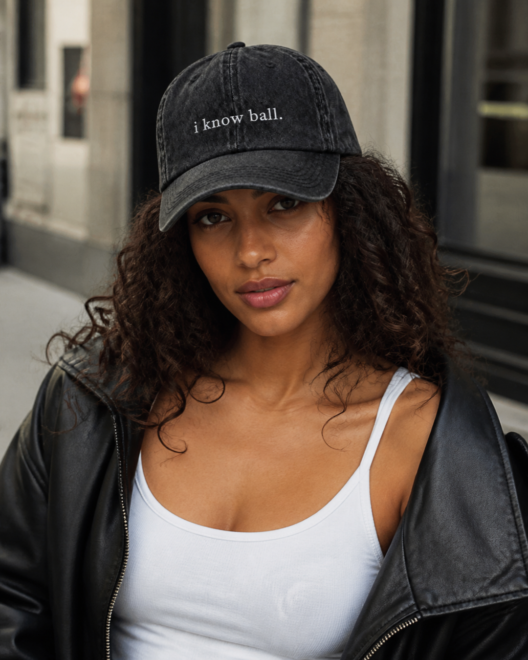 A woman in a black leather jacket and white tank top wearing the washed black 'i know ball.' cap on a city street.