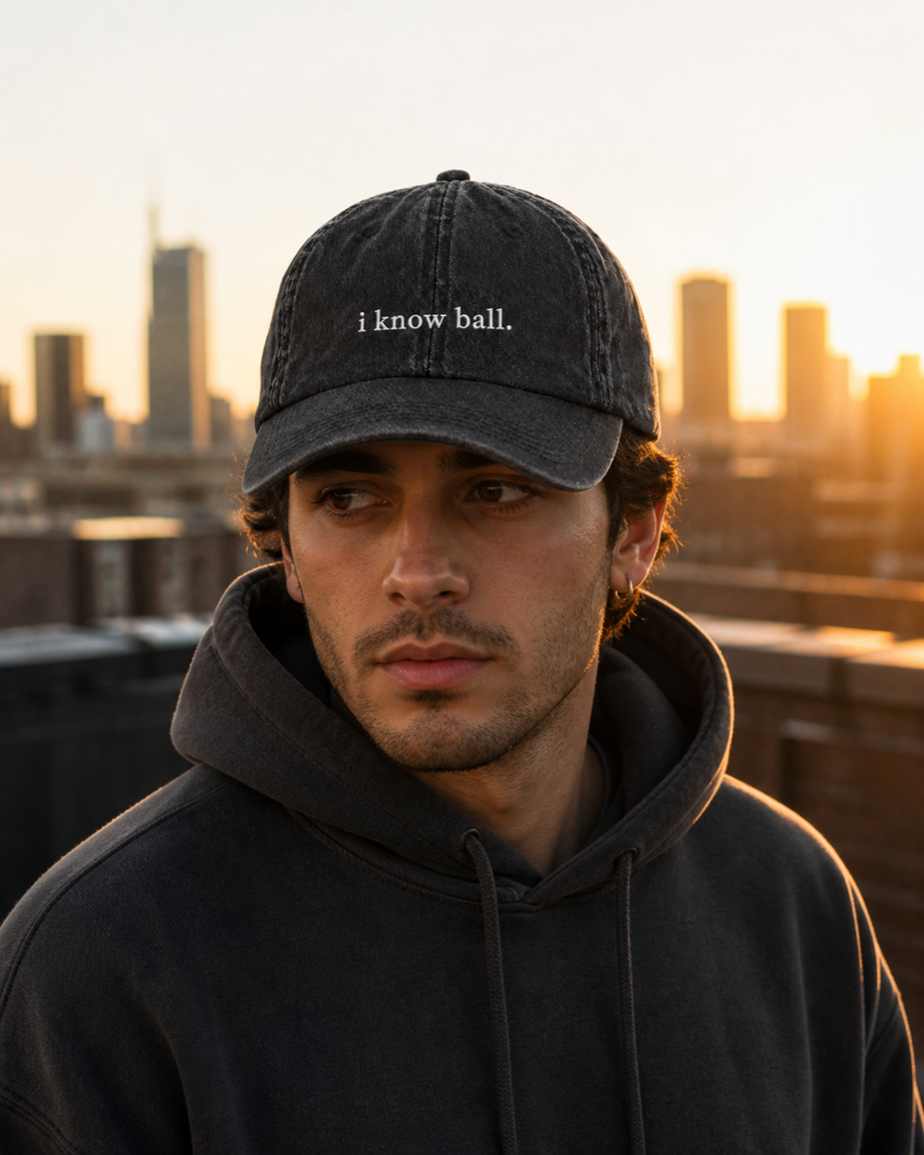 A man in a dark hooded sweatshirt wearing the washed black 'i know ball.' cap on a rooftop at sunset, city skyline behind him.