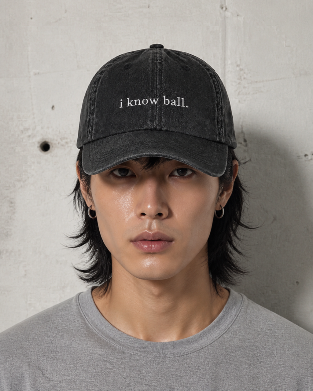 A person in a grey t-shirt wearing the washed black 'i know ball.' cap against a bare concrete wall.