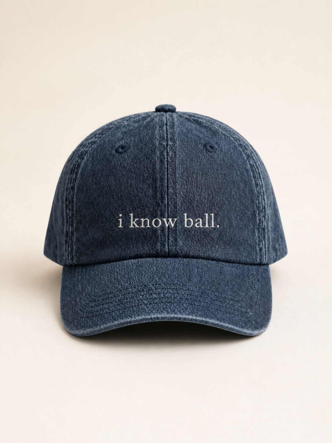 Washed navy 'i know ball.' cap photographed face-on against a warm cream studio backdrop.