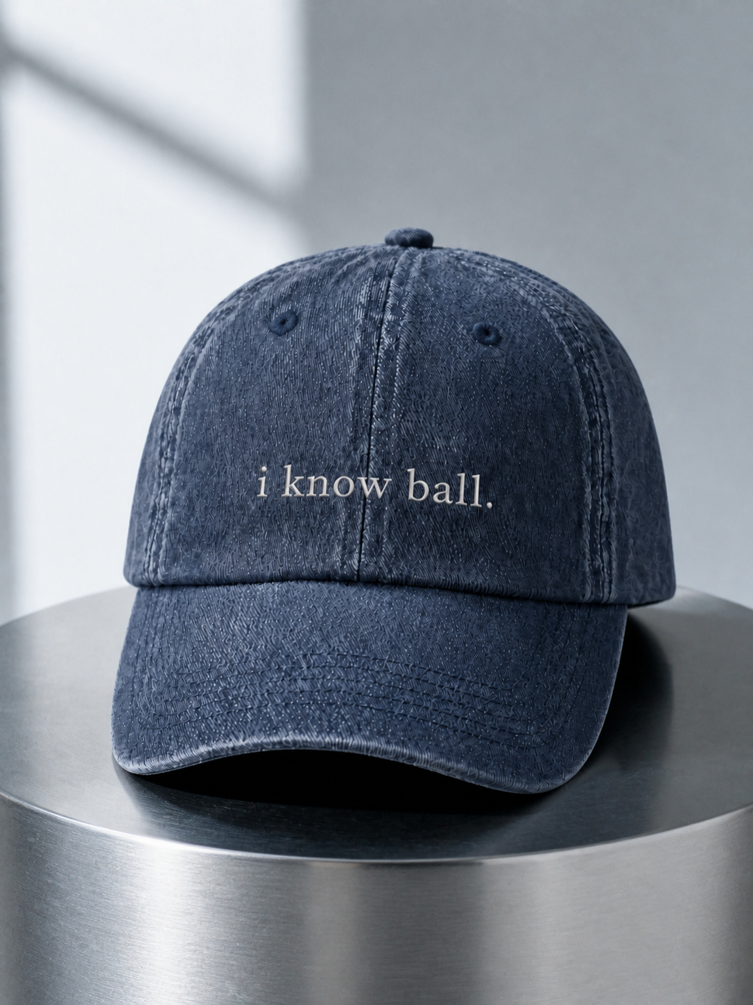 Washed navy 'i know ball.' cap displayed on a polished steel cylinder plinth in cool daylight.