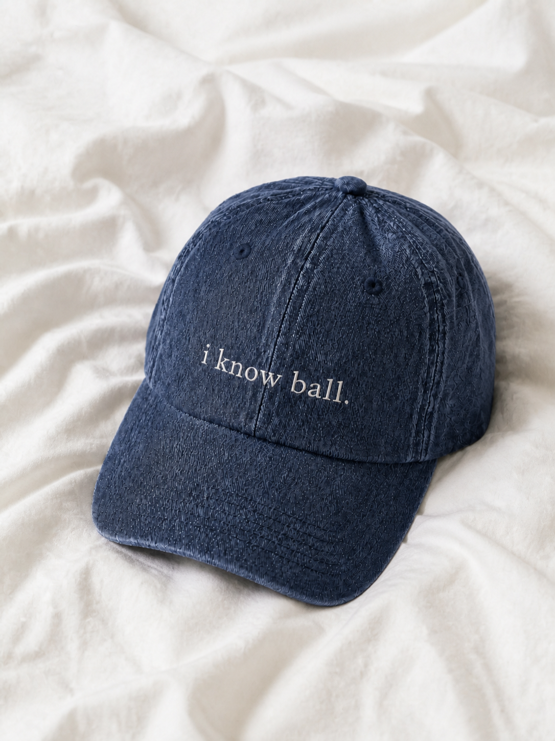 Washed navy 'i know ball.' cap resting on rumpled cream linen.