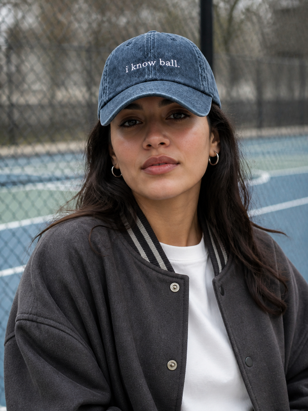 A woman in a charcoal sweatshirt wearing the washed navy 'i know ball.' cap on a city street in front of a stone building.