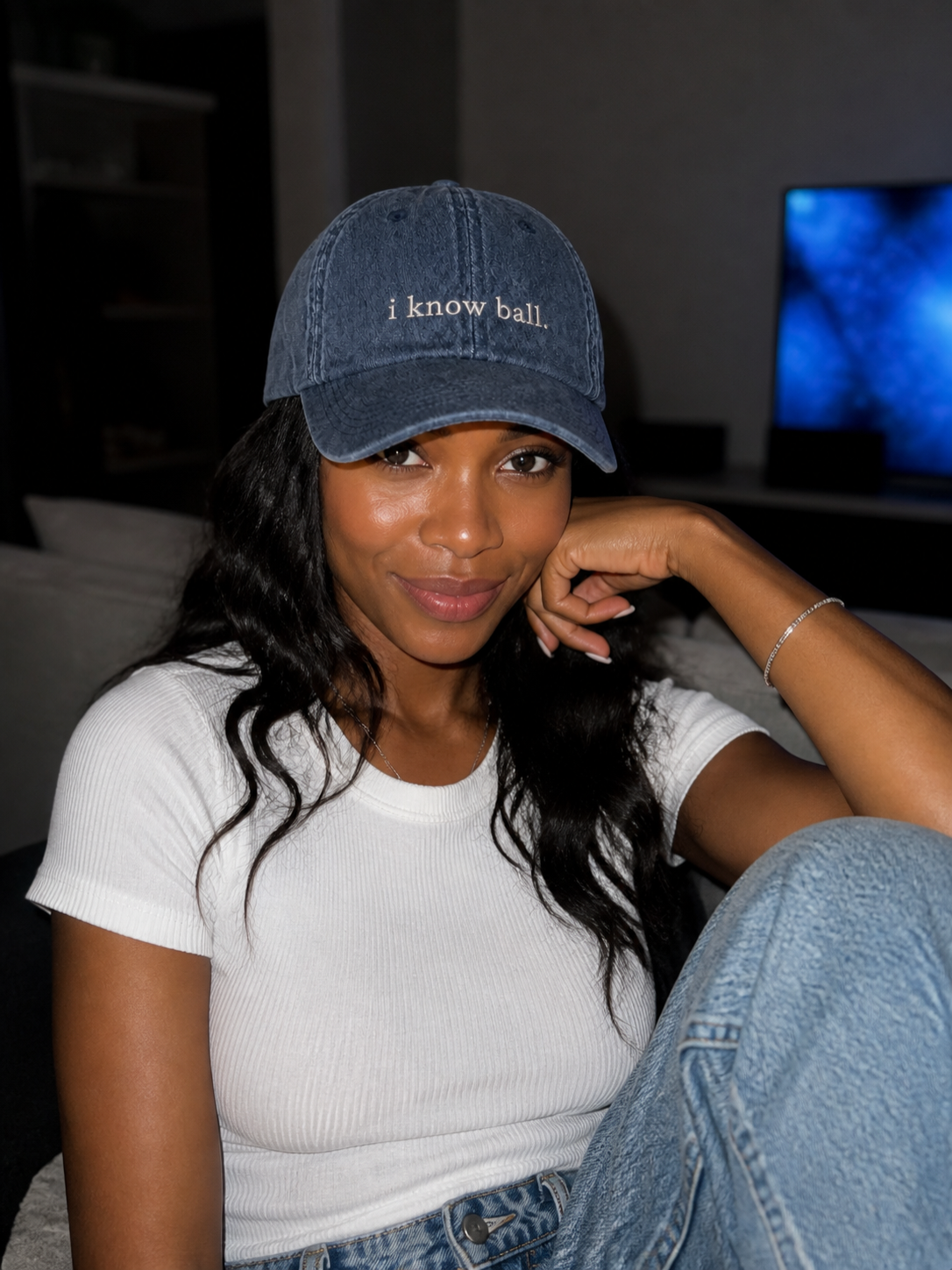 A woman in a white tank top wearing the washed navy 'i know ball.' cap, seated indoors by a window at dusk.