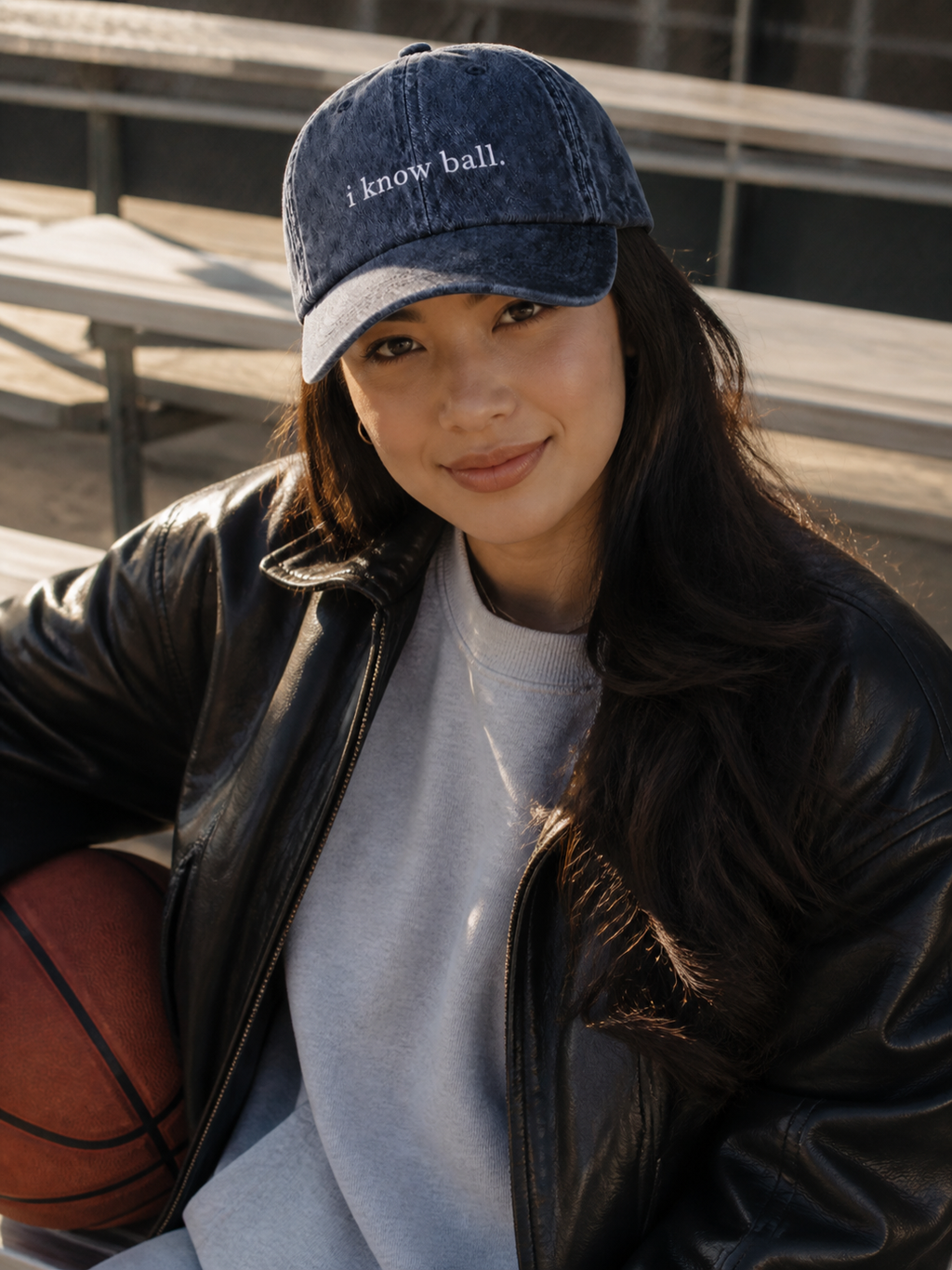 A woman in a black leather jacket holding a coffee cup and wearing the washed navy 'i know ball.' cap at a cafe terrace.
