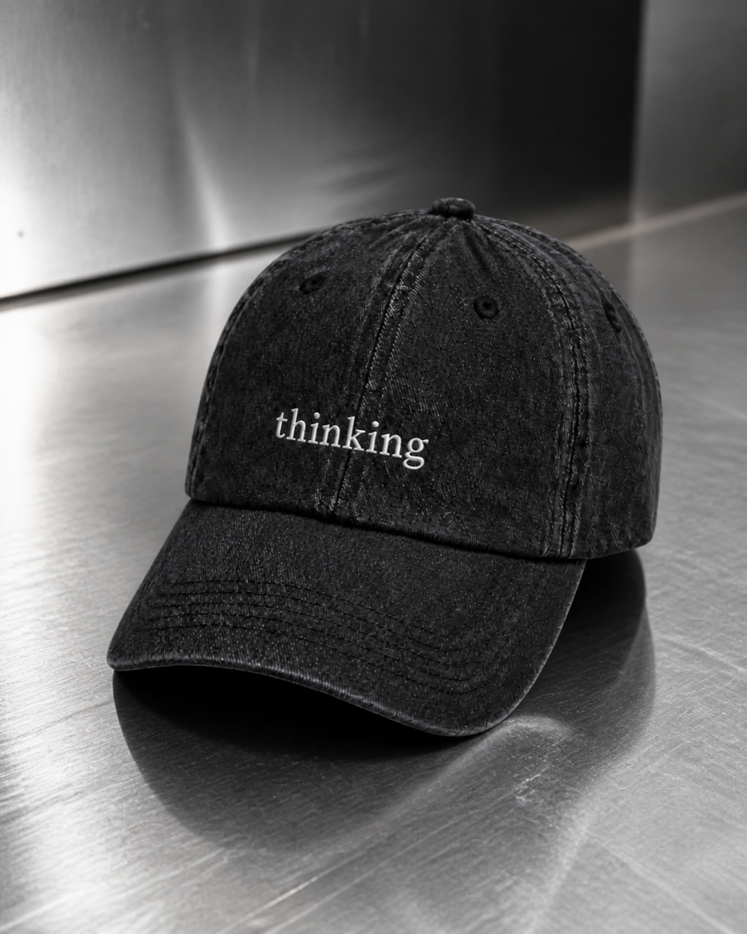 Washed black 'thinking' cap resting on a brushed steel surface under cool directional light.