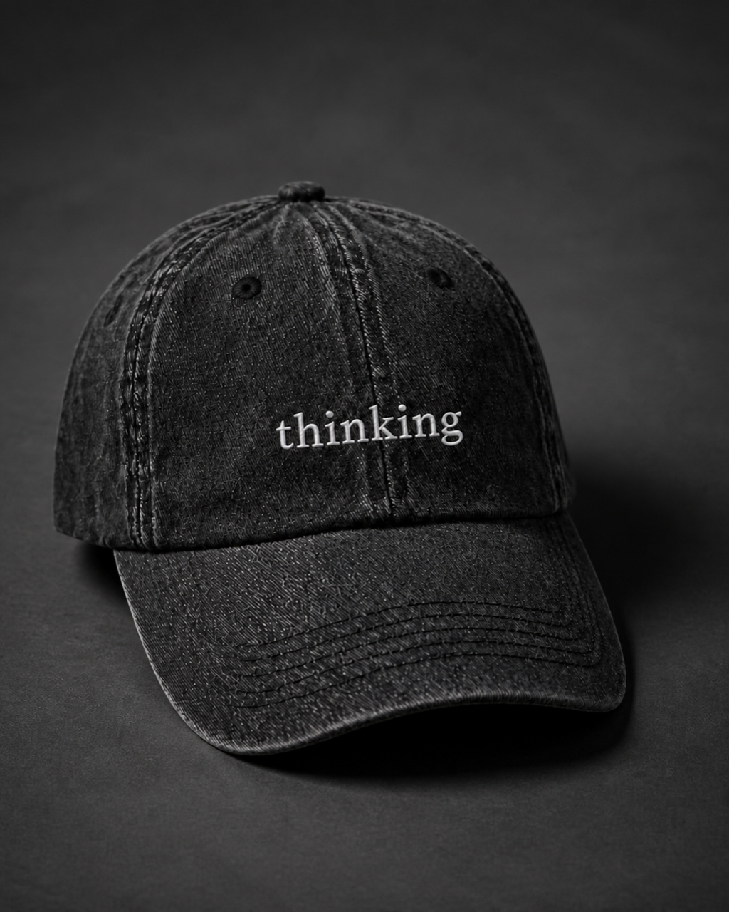 Washed black 'thinking' cap on a dark charcoal backdrop, lit from the side to pick out the embroidery.