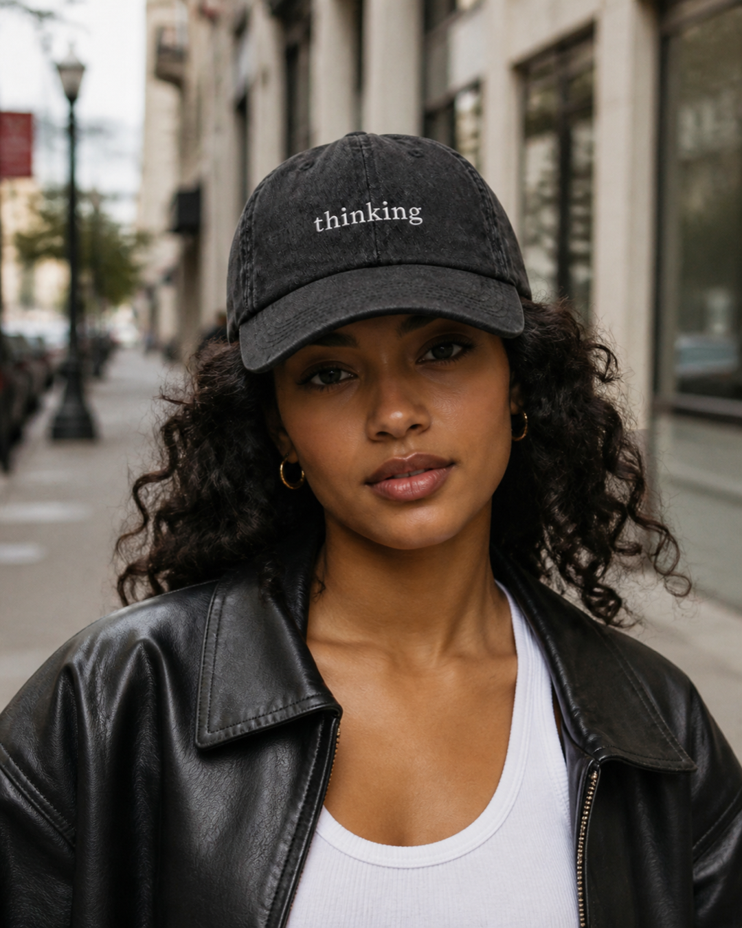 A woman in a black leather jacket and white tank top wearing the washed black 'thinking' cap on a city street.