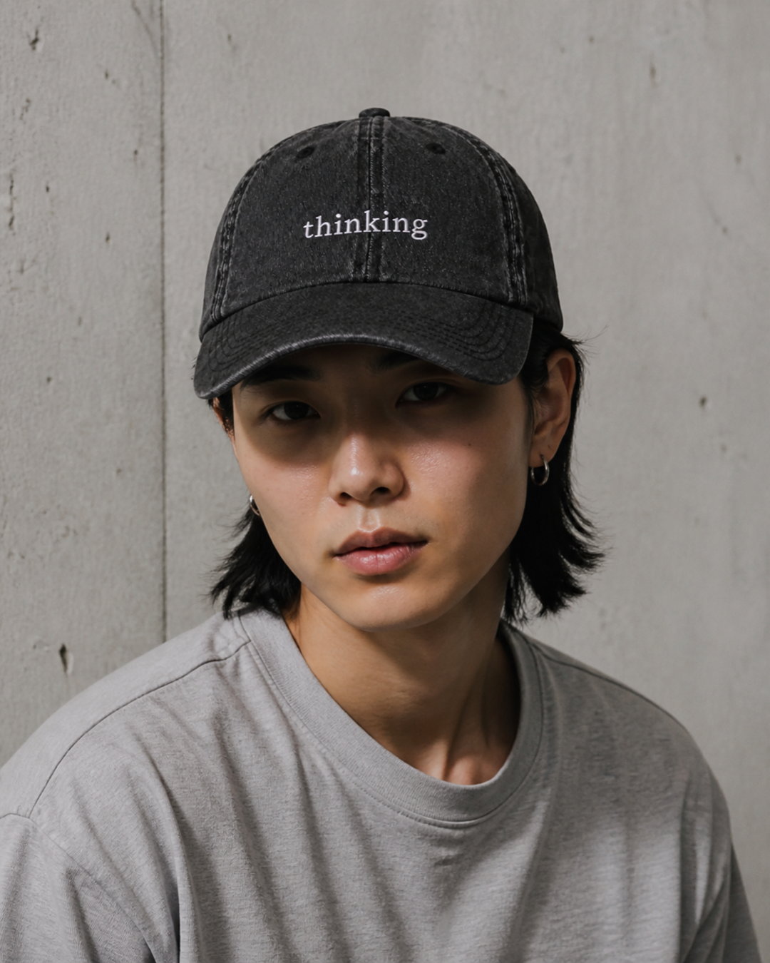 A person in a grey t-shirt wearing the washed black 'thinking' cap against a bare concrete wall.