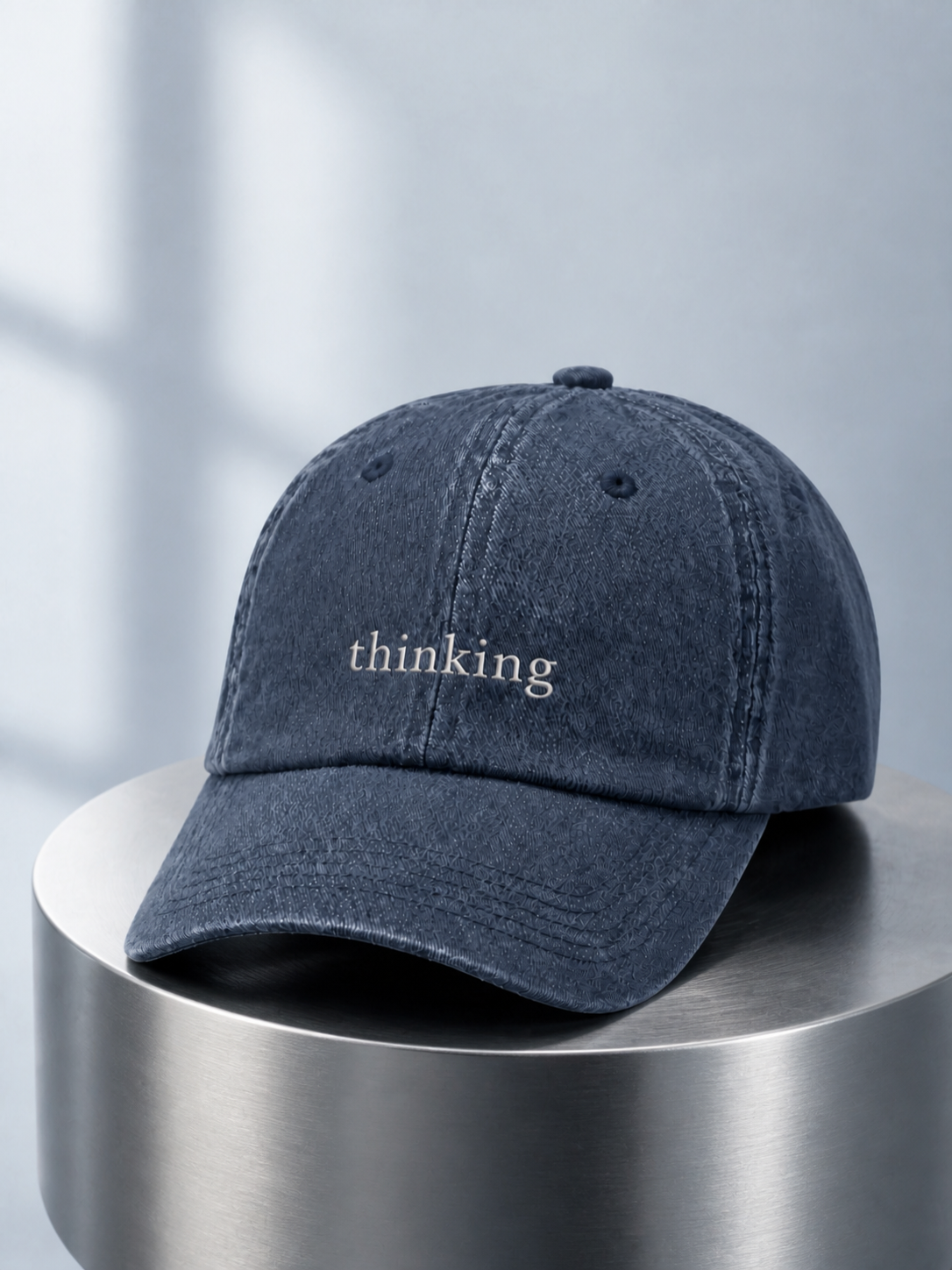 Washed navy 'thinking' cap displayed on a polished steel cylinder plinth in cool daylight.